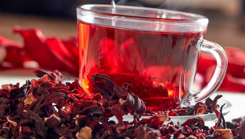Mesa com hibiscos e uma xicara de vidro com o chá de hibisco.