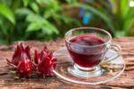 Mesa de madeira com hibiscos e uma xicara de vidro com chá de hibisco.