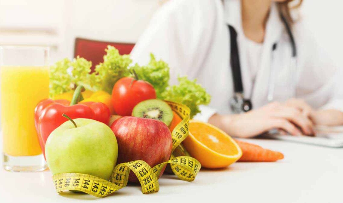 Sala de uma nutricionista que está sentada em sua mesa com seus equipamento e alimentos saudáveis.