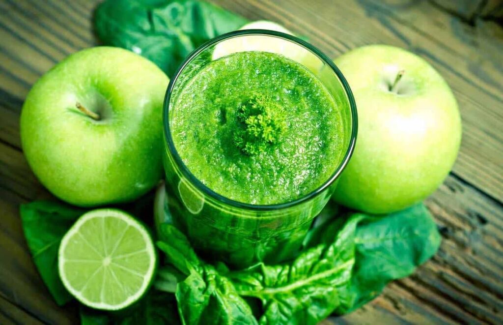 Mesa posta com uma jarra de vidro ao centro com o suco verde em volta com maçãs verdes, limão e espinafre.