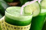 Dois copos de vidro com suco verde, canudos e uma rodela de pepino em cada, uma fita métrica em volta do copo da frente.