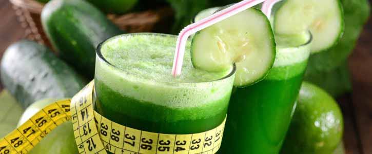 Dois copos de vidro com suco verde, canudos e uma rodela de pepino em cada, uma fita métrica em volta do copo da frente.