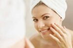 Mulher com toalha de banho nos cabelos, analisando sua pele no espelho após utilizar colágeno.