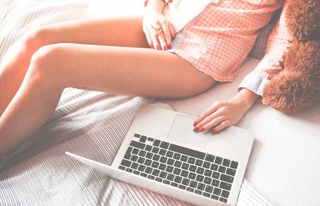 Mulher deitada na cama com uma camiseta xadrez e com o seu notebook do lado.