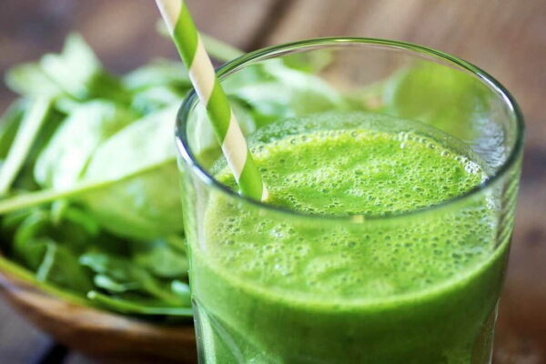 Mesa com um copo de vidro com suco verde detox e uma tigela de folhas verdes ao fundo.