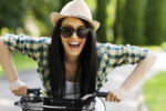Mulher sorrindo andando de bicicleta ao ar livre com um chápeu na cabeça.