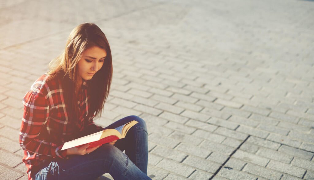 Mulher sentada em uma calçada lendo um livro vermelho.