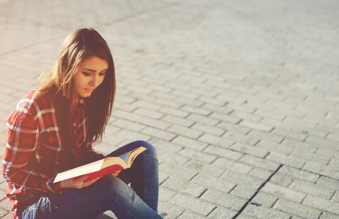 Mulher sentada em uma calçada lendo um livro vermelho.