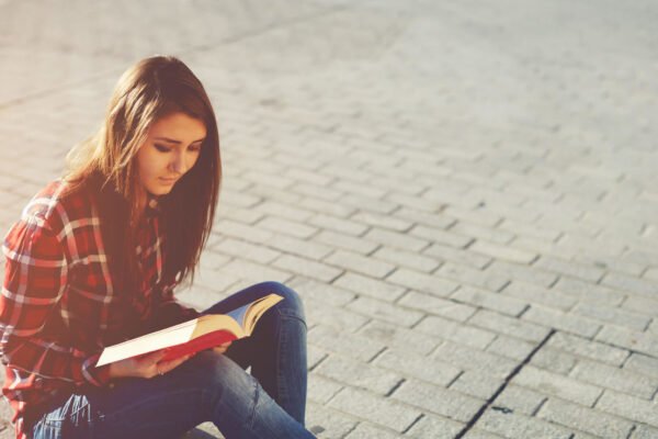Mulher sentada em uma calçada lendo um livro vermelho.