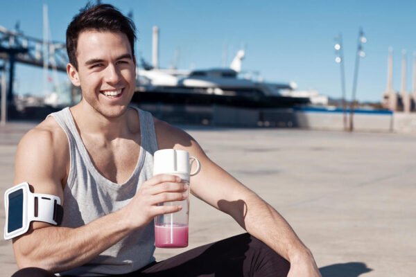 Homem sorrindo sentado em um estacionamento com roupas de treino e segurando uma garrafa com suplemento.