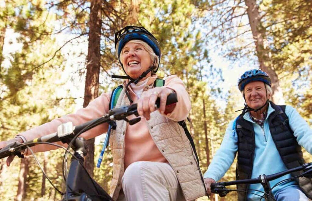 Um casal de idosos se divertindo andando juntos de bicicleta com capacetes em uma rua arborizada.