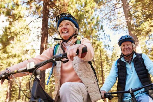 Um casal de idosos se divertindo andando juntos de bicicleta com capacetes em uma rua arborizada.