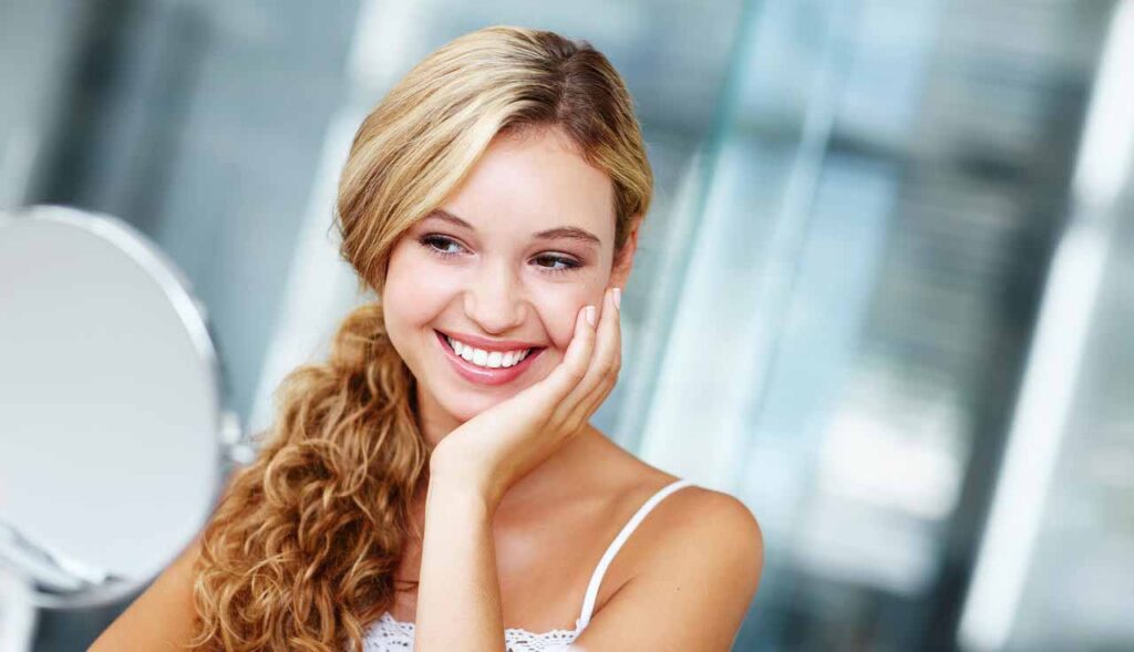 Mulher sorrindo se olhando em um espelho de mão enquanto toca seu rosto.