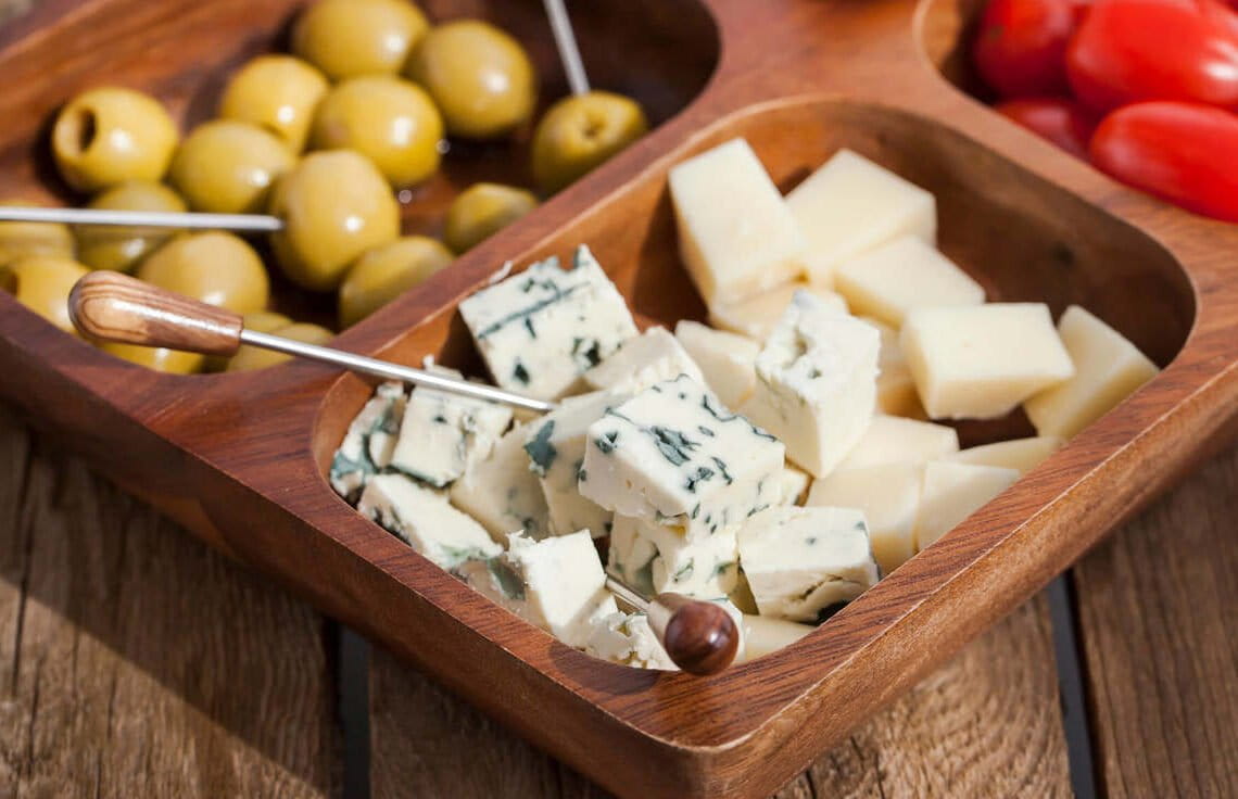 Mesa com prato de madeira com divisórias com opções de petiscos saudáveis, como queijos, azeitonas e tomates cereja.