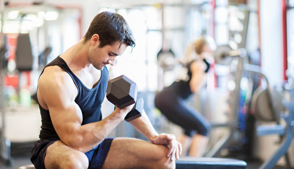 Homem em uma academia sentado em um banco enquanto faz exercício de braço unilateral com um halter.