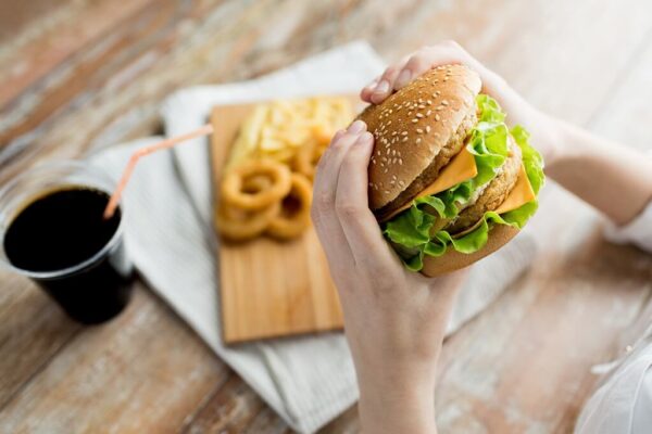 Mulher sentada em uma mesa comendo hambúrguer, batata frita, onion rings e bebendo refrigerante.