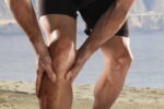 Homem em uma praia com as mãos em um dos joelhos, representando dores nas articulações.