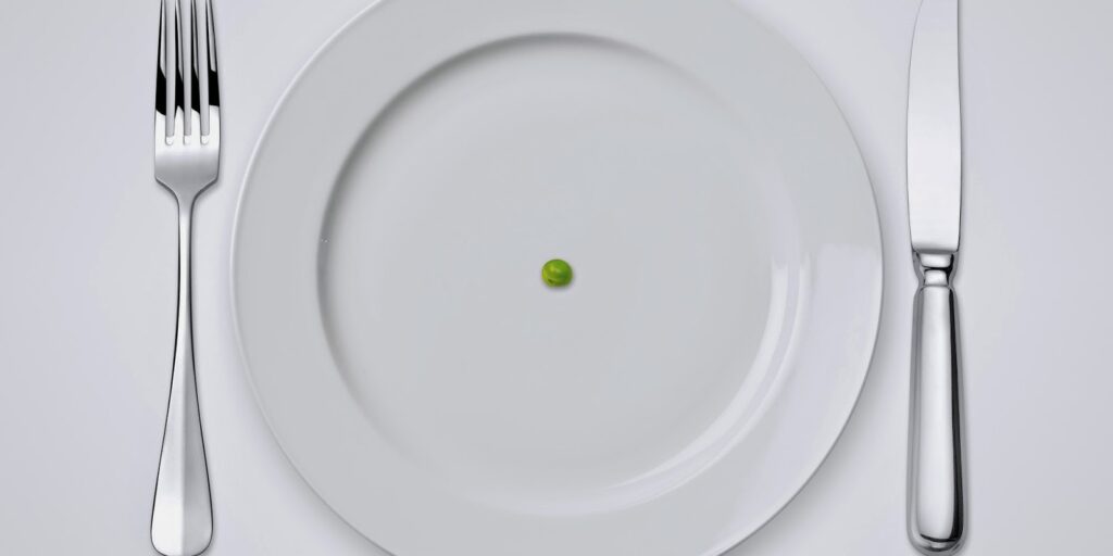 Mesa com um garfo e uma faca ao lado de um prato branco com uma única ervilha no centro.