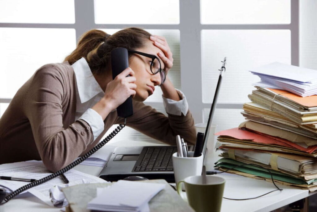 Mulher estressada em seu serviço falando ao telefone enquanto olha para o seu notebook na mesa.