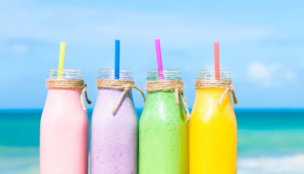 Quatro garrafas de vidro com shakes e canudos de cores diferentes em cada e uma praia ao fundo.