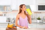 Mulher na cozinha de sua casa, tomando sua proteina em uma garrafa amarela.