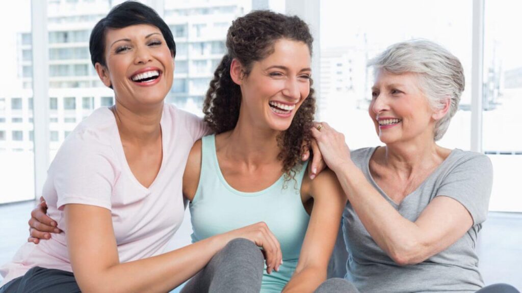 Três mulheres de diferentes gerações e idades, sentadas se abraçando e sorrindo.
