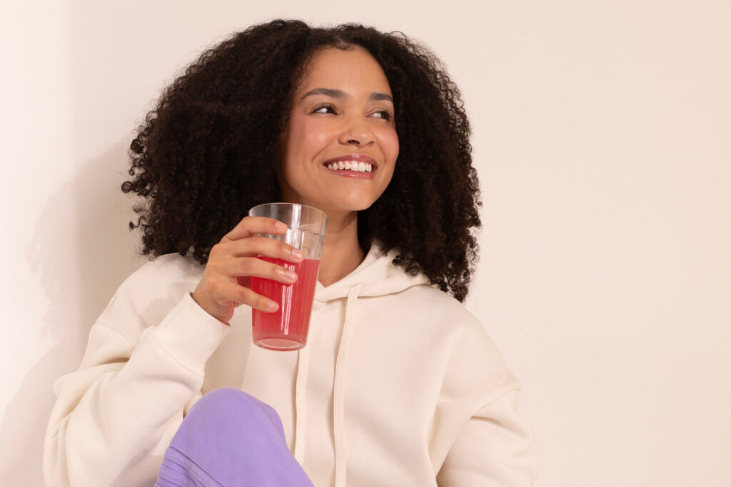 Mulher sorrindo enquanto toma um copo de colágeno em pó diluído em bebida vermelha.
