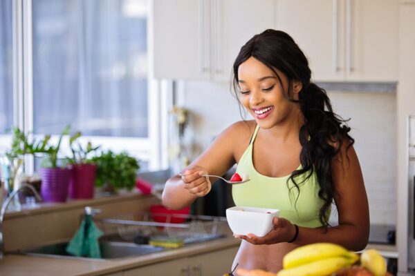Mulher sorrindo em sua casa enquanto faz uma refeição com uma tigela branca nas mãos.