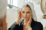 Mulher com toalha de banho nos cabelos, se olhando no espelho com as mãos no rosto, após tomar seu colágeno.