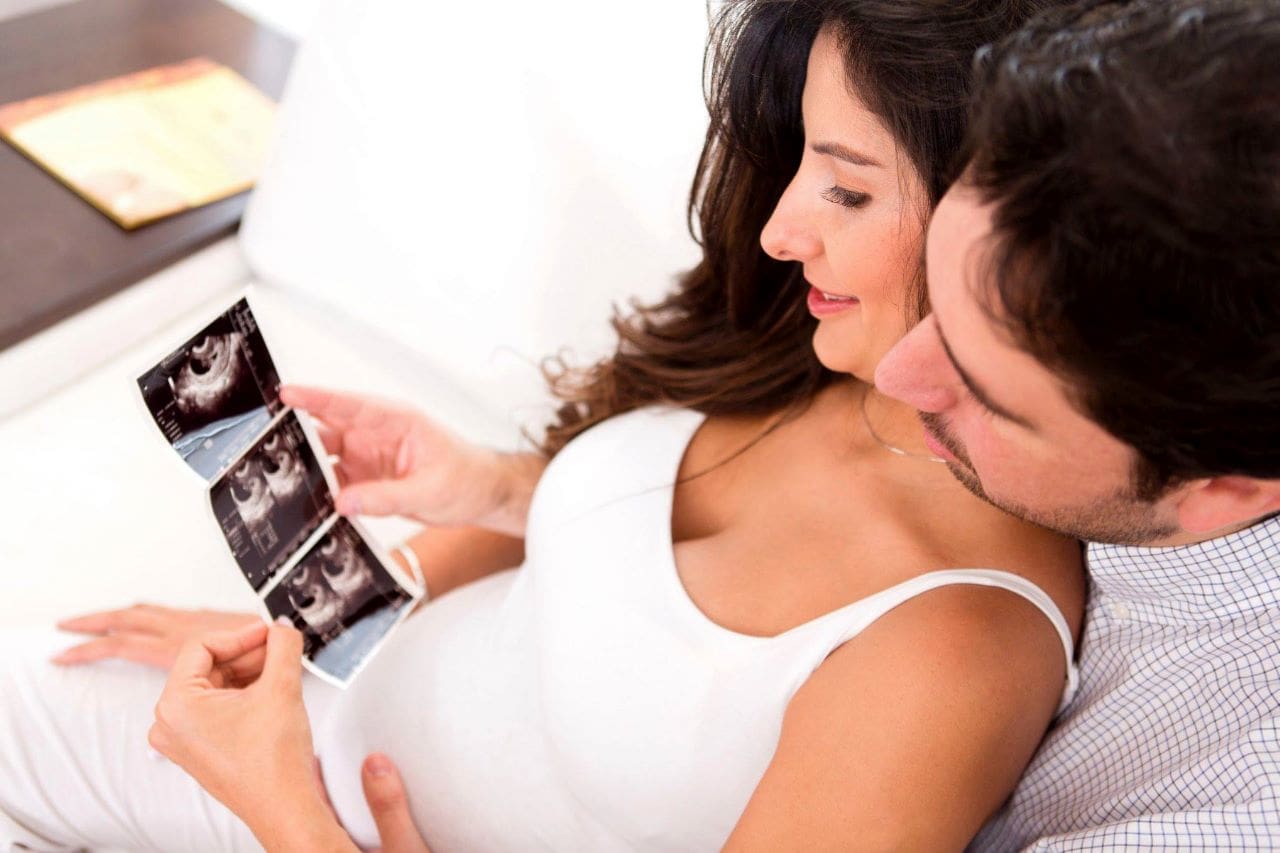 Homem e mulher grávida, sentados e abraçados, vendo fotos de uma ultrassom 