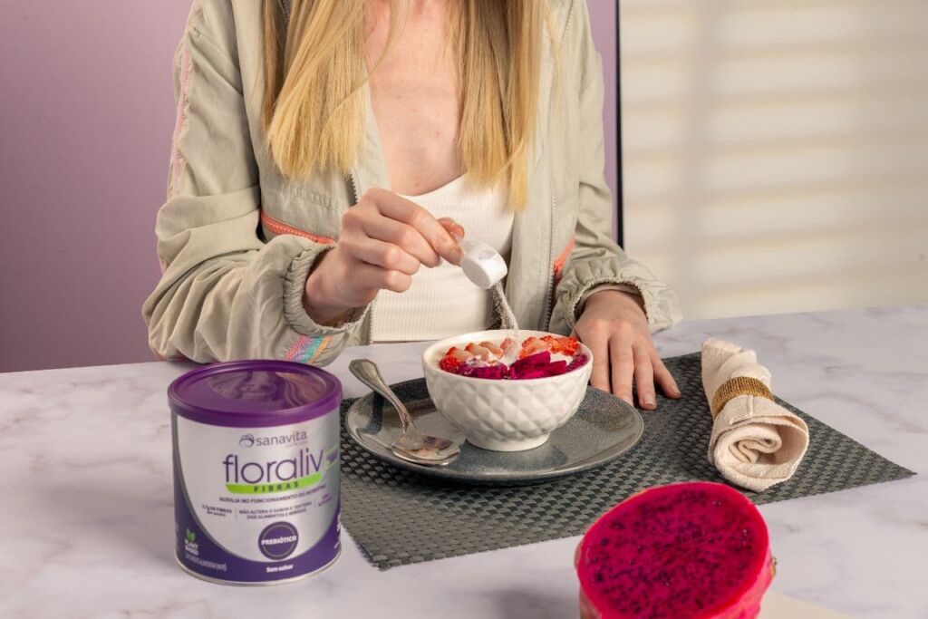 Mulher adicionando uma dose de Probiótico Floraliv Sanavita em uma tigela com iorgute e frutas 
