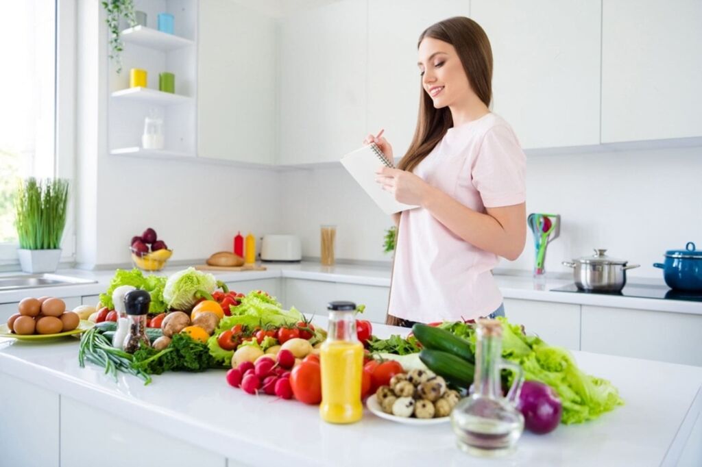 Mulher na cozinha fazendo anotações em um caderno, com uma bancada repleta de saladas, frutas e verduras  