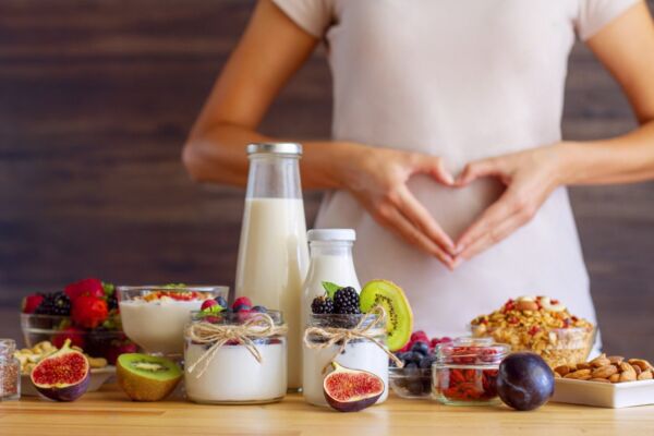 Mesa com alimentos ricos em probióticos, como leite e iogurtes e mulher ao fundo com as mãos em forma de coração na barriga