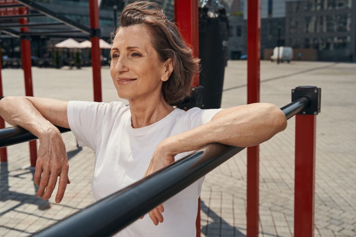 Mulher de meia idade, sorrindo, com os braços apoiados em barras de metal para exercício no parque.