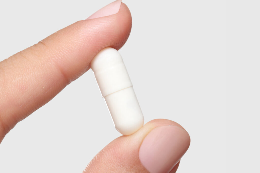 Mão segurando cápsula de resveratrol entre os dedos, em fundo cinza claro.