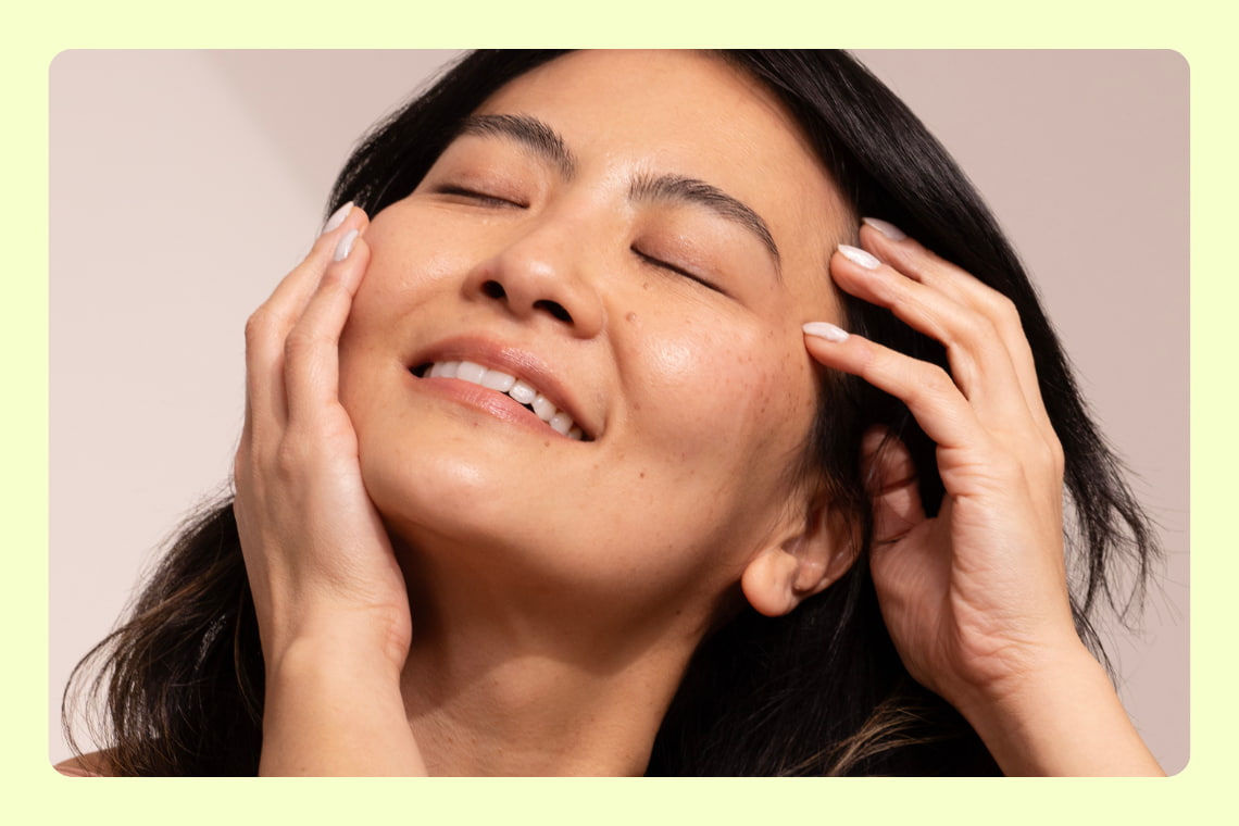 Mulher sorridente com olhos fechados toca o rosto, transmitindo bem-estar e autocuidado.