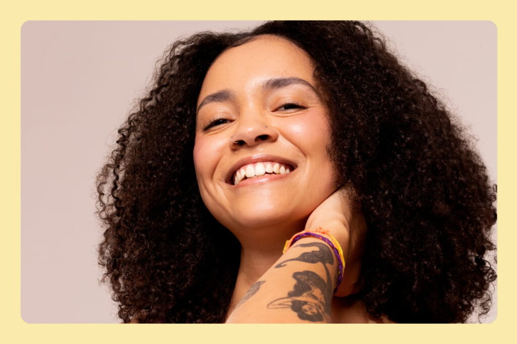Mulher sorridente com cabelo cacheado, piercing no nariz e tatuagem no braço, usando pulseiras coloridas, em fundo neutro.