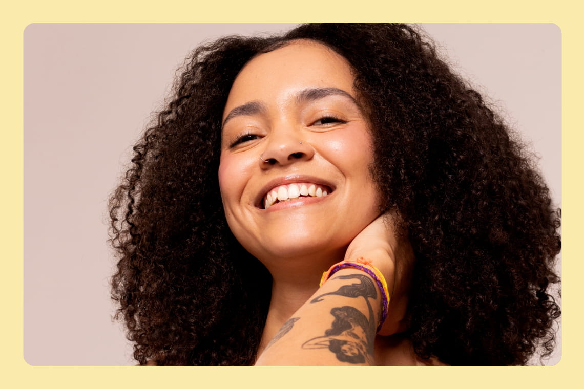 Mulher sorridente com cabelo cacheado, piercing no nariz e tatuagem no braço, usando pulseiras coloridas, em fundo neutro.