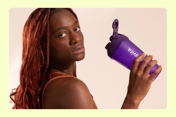 Mulher com tranças vermelhas segurando coqueteleira roxa de suplemento esportivo em fundo neutro.