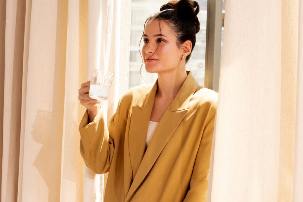 Mulher em pé próxima à janela, segurando copo com creatina, iluminada por luz natural, em ambiente interno aconchegante.