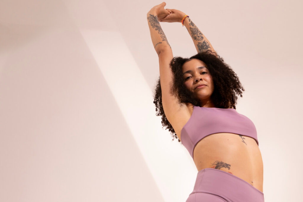 Mulher com roupas fitness lilás alongando os braços acima da cabeça, exibindo tatuagens, em ambiente interno com fundo neutro.